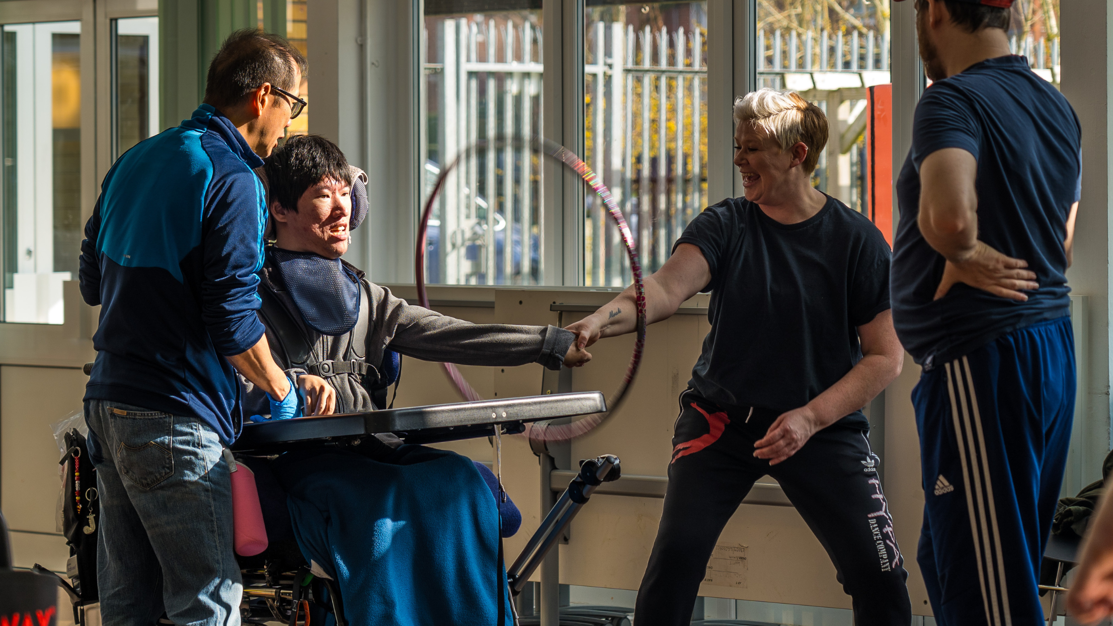 The image shows a community creative dance session in progress. Two staff members, one wearing blue, are assisting participants with disabilities. One staff member is guiding a person in a wheelchair through dance movements, both smiling with engagement. Another participant dances freely with expressive body movements nearby. The setting is an indoor facility with large windows, appearing to be a space designed for inclusive artistic expression through dance for individuals of all abilities.