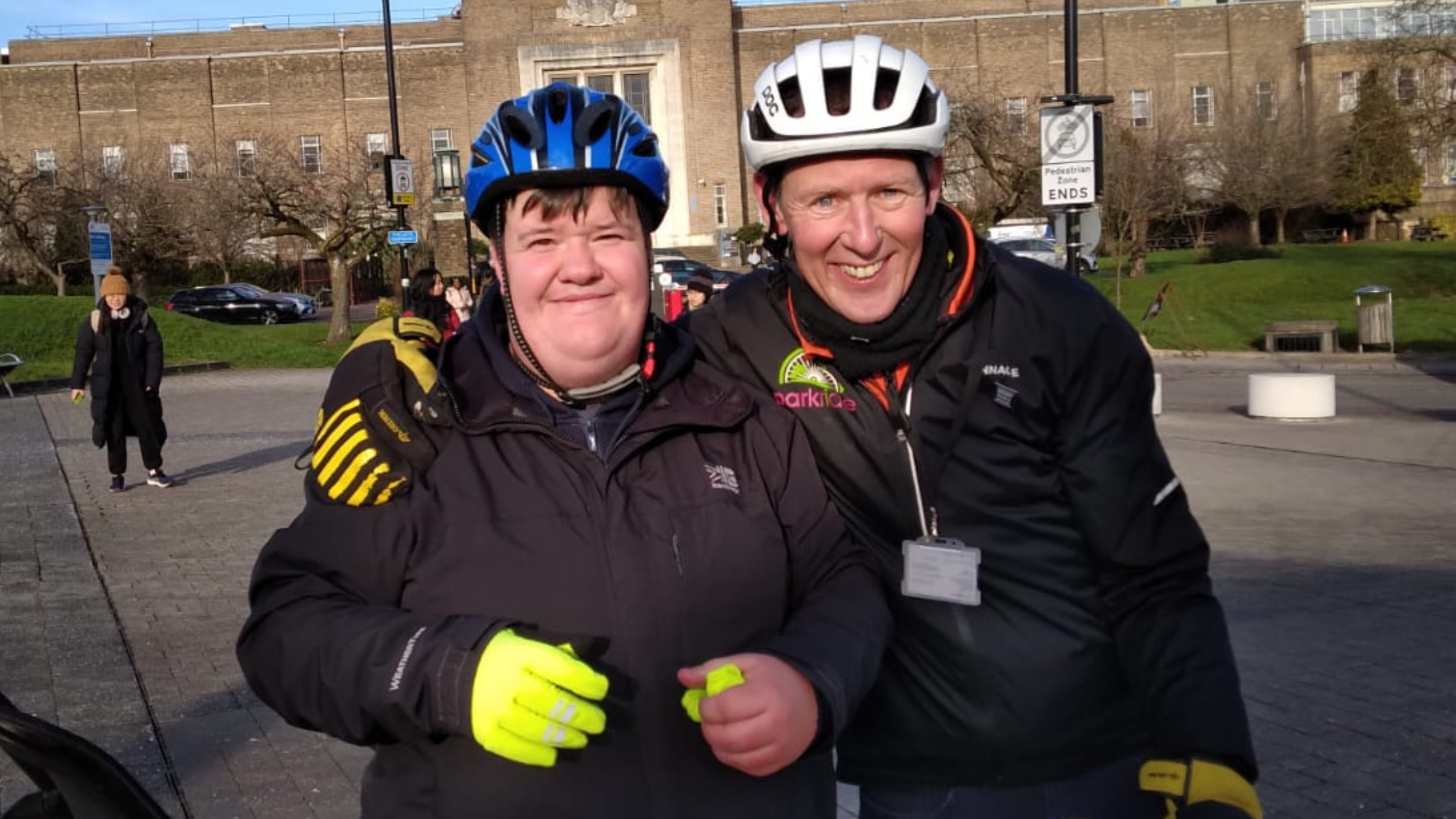 The image shows two cyclists, likely friends, standing together outside on what appears to be a university campus or similar institutional setting with brick buildings in the background. They are dressed in cycling gear, wearing helmets and reflective jackets, suggesting they have been out for a bike ride. Both individuals have big smiles on their faces, conveying a sense of joy and camaraderie. The scene captures the enjoyment of participating in outdoor recreational activities together.