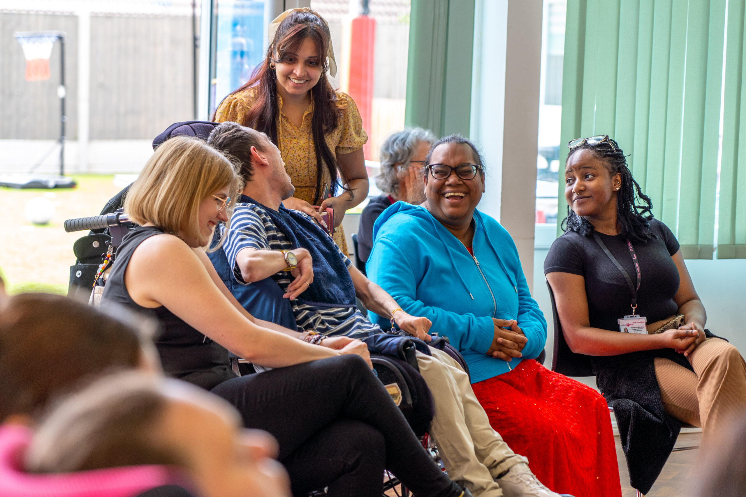 Group of diverse people sitting together indoors, smiling and interacting. Includes people using wheelchairs and mobility aids.