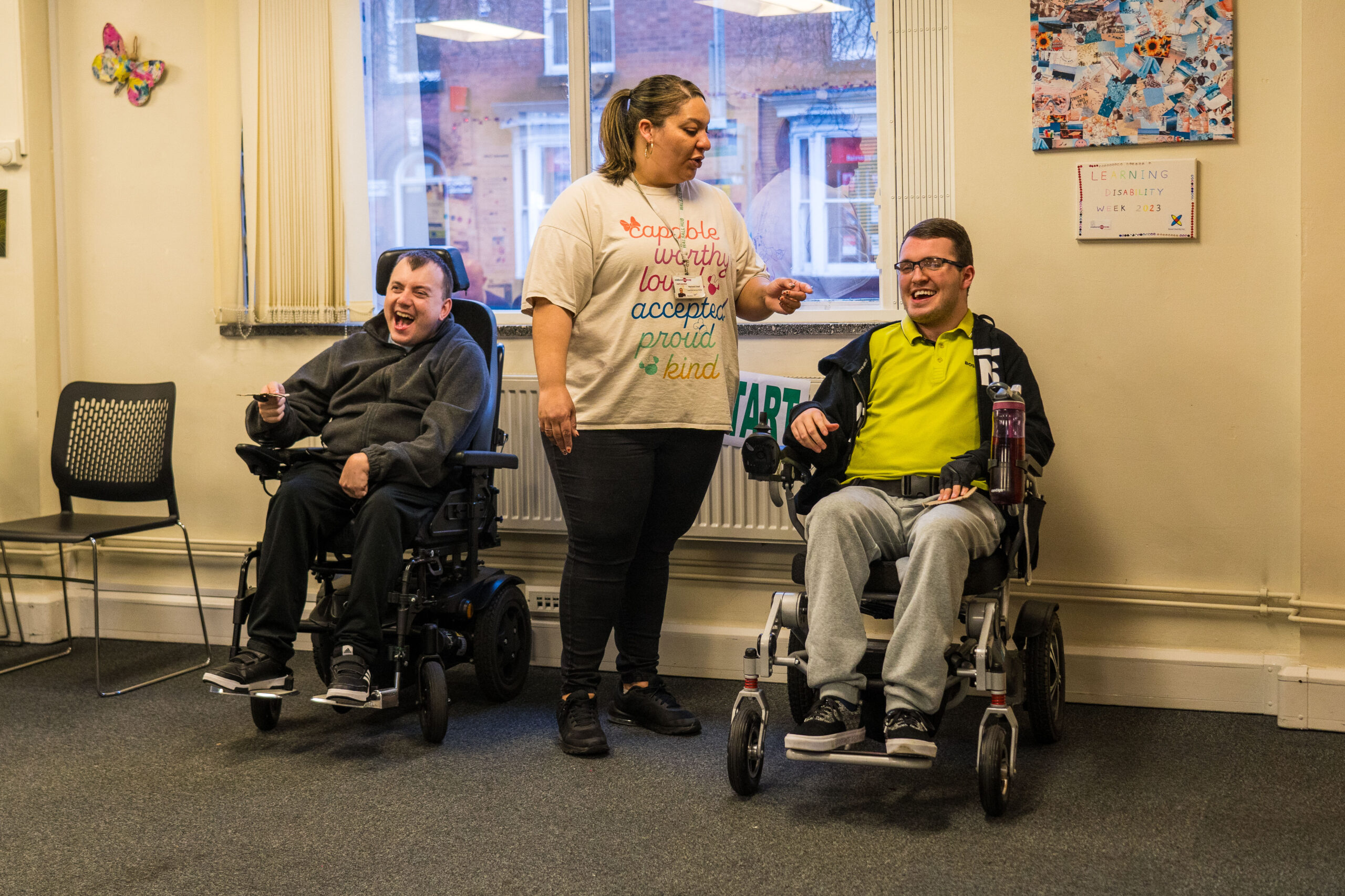 A woman stands between two men in wheelchairs in a community center room. The woman wears a t-shirt with positive words like 'capable', 'worthy', and 'kind'. The men are smiling and appear to be engaged in conversation. The room has colorful decorations including a butterfly on the wall and a poster about learning disability week.