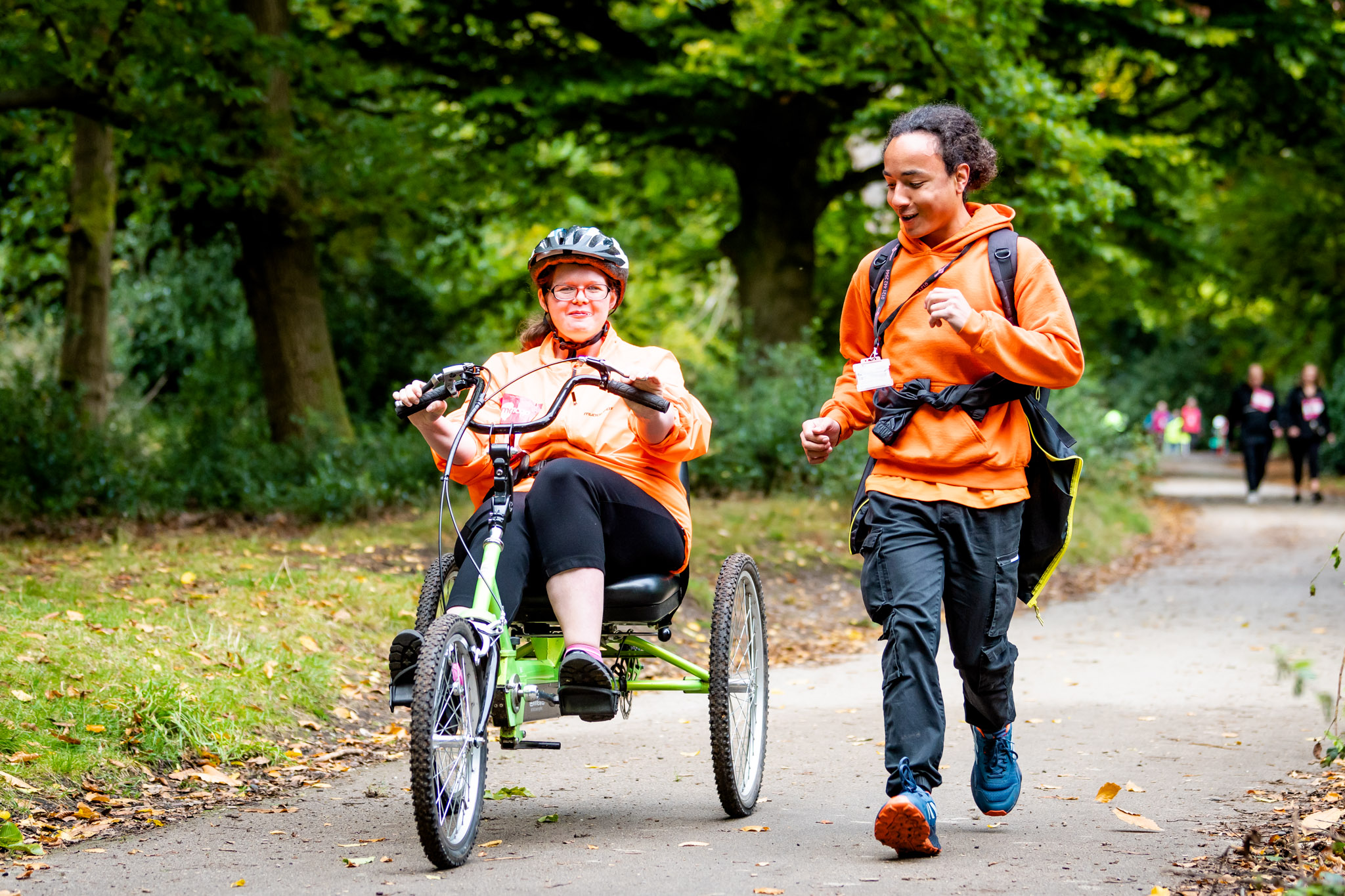 A woman riding an accessible tricycle alongside a support worker in a forest path. Both wear orange sweatshirts, and the woman focuses on cycling while the worker runs beside her.