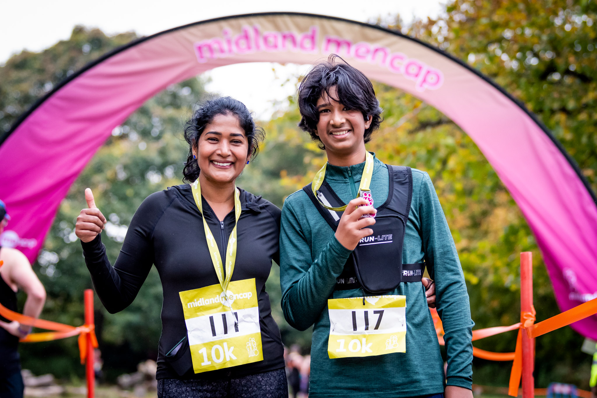 A smiling mother and son, both with medals around their necks, celebrate after completing a 10K race. They stand in front of a Midland Mencap arch, proud of their achievement.
