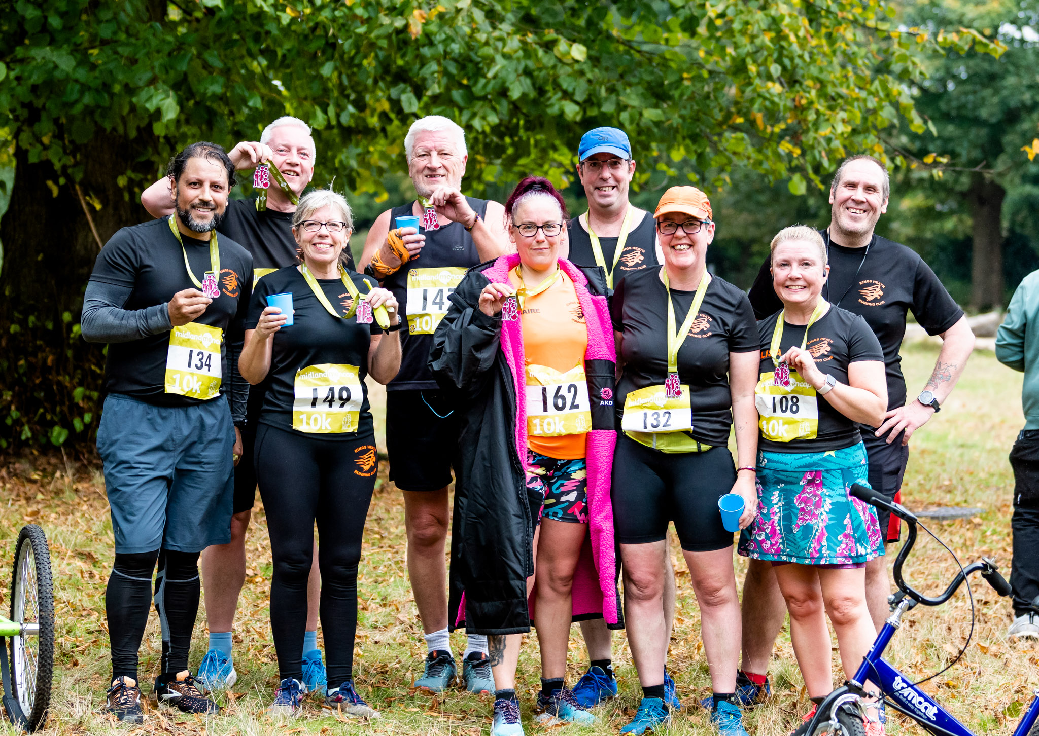 A group of eight smiling runners, all wearing 10K race bibs, proudly show off their medals after completing the race. They stand together in a park, some holding drinks, dressed in running gear with bright accents. The group looks jubilant as they celebrate their accomplishment, surrounded by trees and nature. A tricycle is partially visible on the left side of the image.