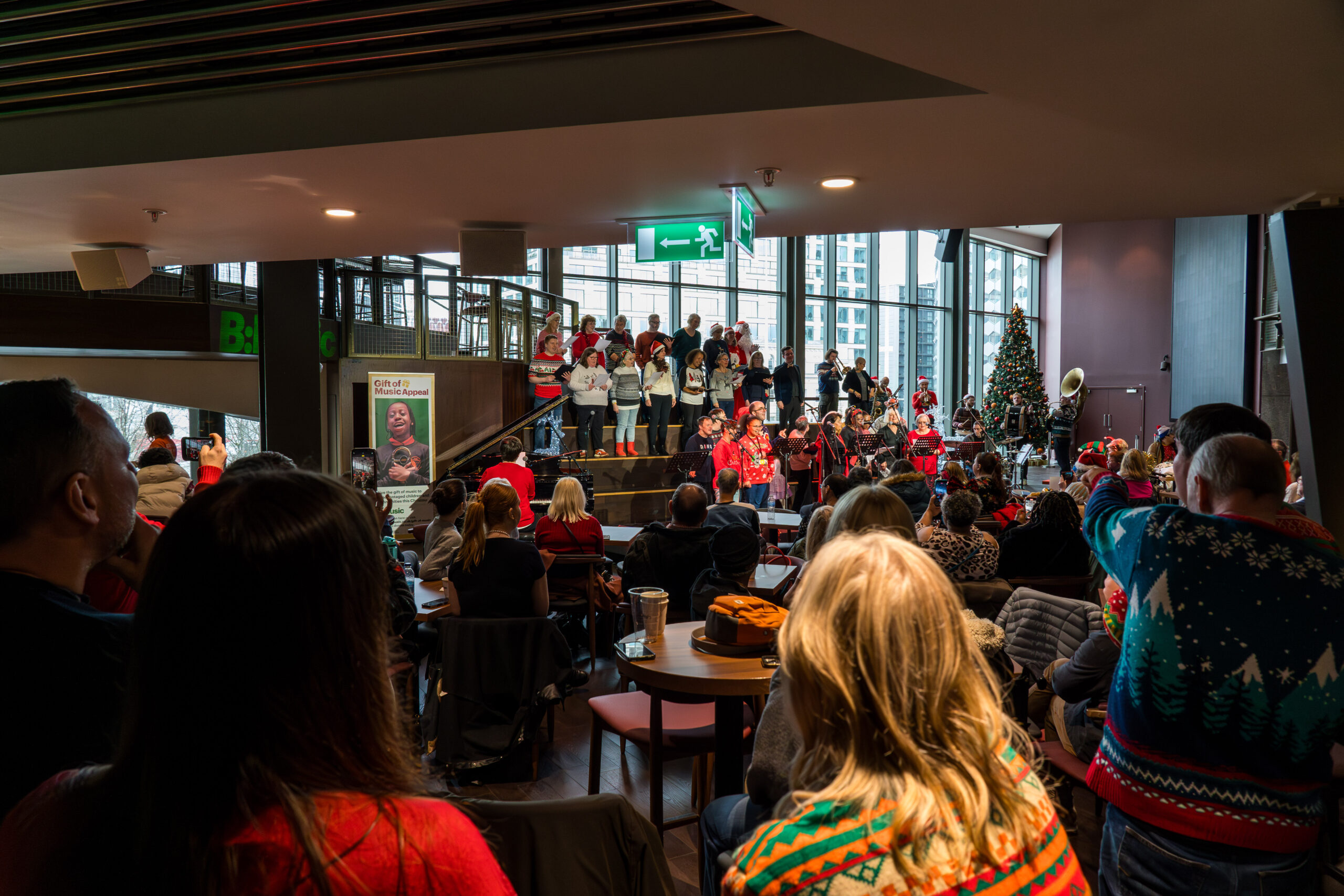 A large audience enjoys a holiday performance featuring a choir and brass band on a tiered stage, framed by festive decorations, a Christmas tree, and city views.