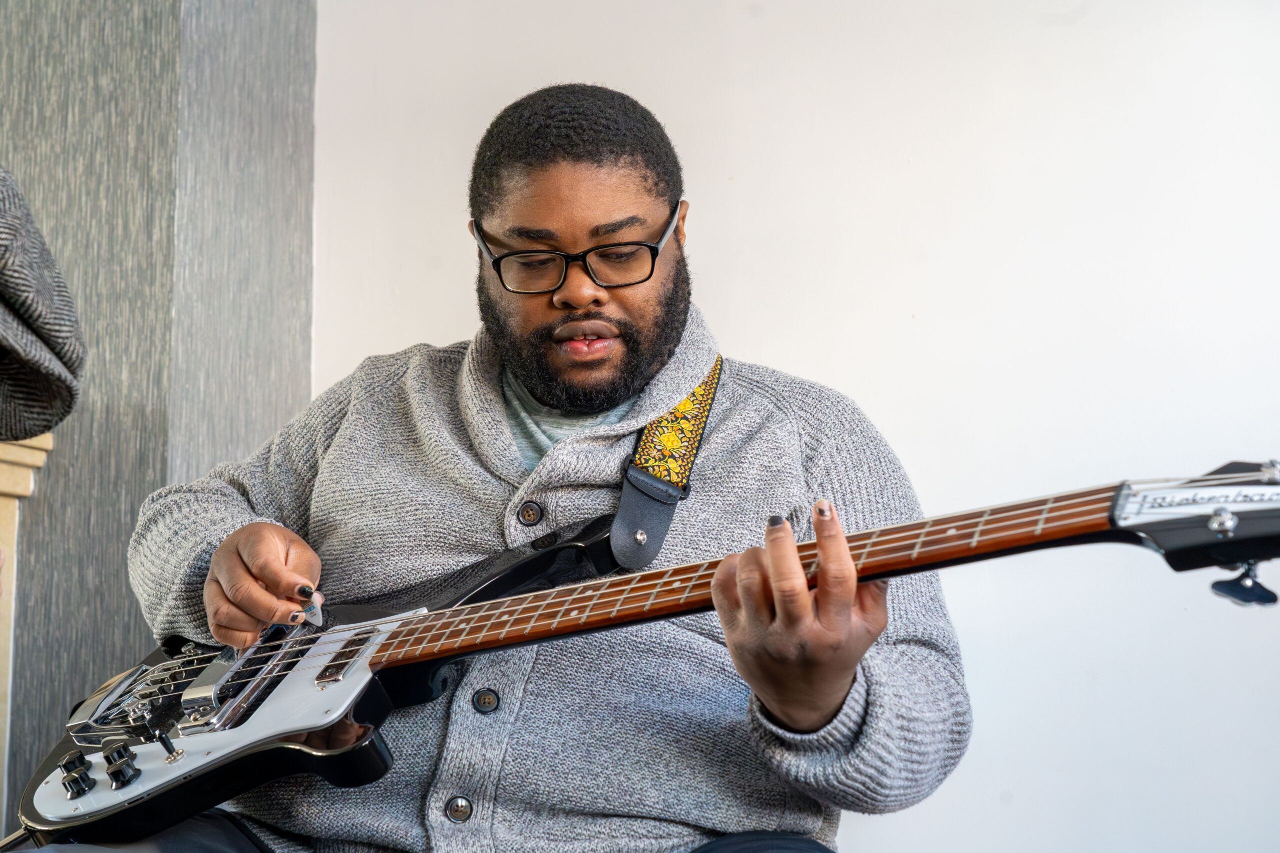 A man wearing glasses and a grey cardigan is playing a black and white electric bass guitar. He is looking at the instrument, plucking the strings with his right hand and pressing the frets with his left hand. The background is a simple white and grey wall.