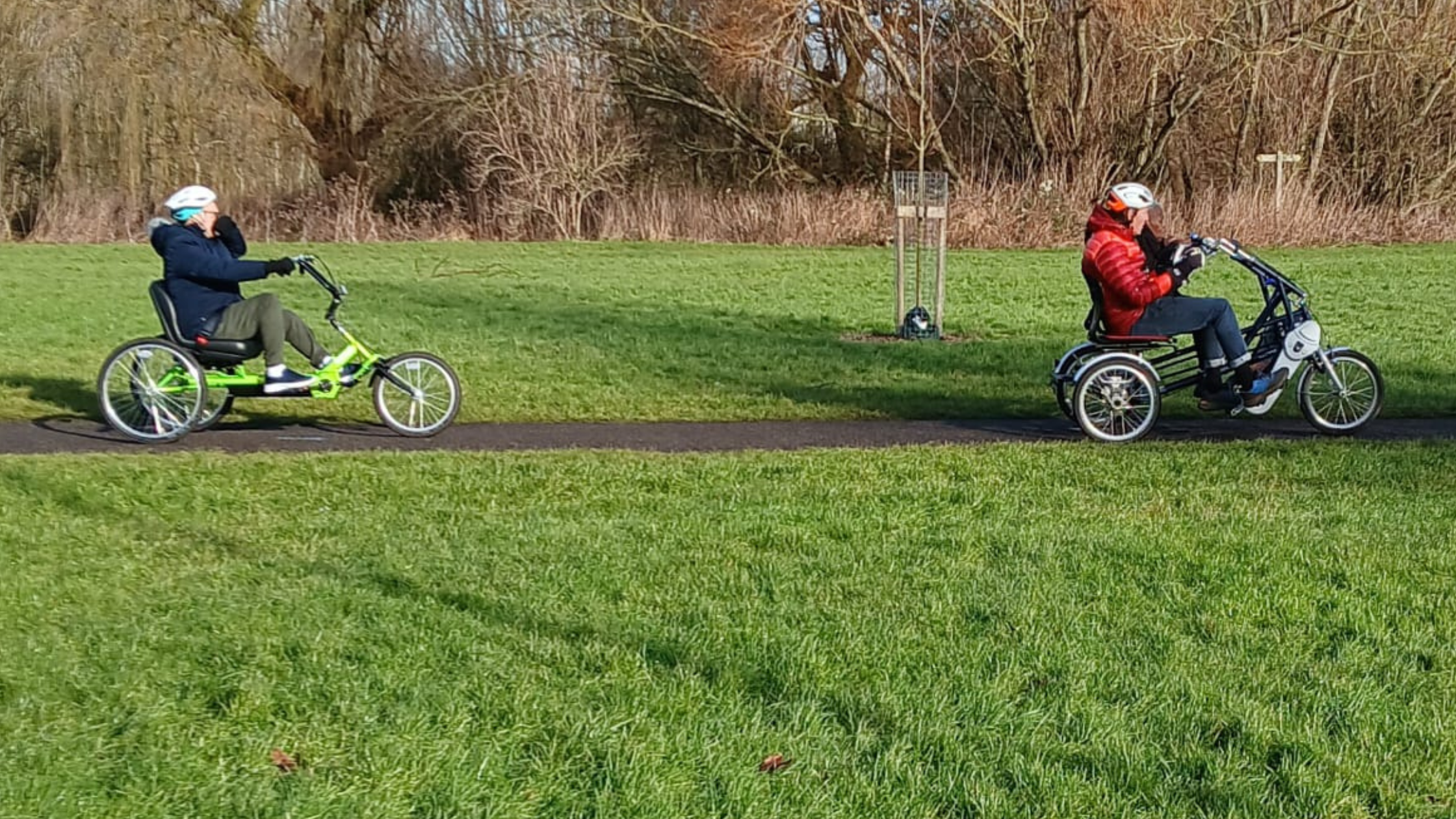 Two cyclists ride adaptive tricycles on a paved path in a grassy park. One cyclist on the left rides a green tricycle, while the other, in a red jacket, rides a larger tandem tricycle with a passenger.