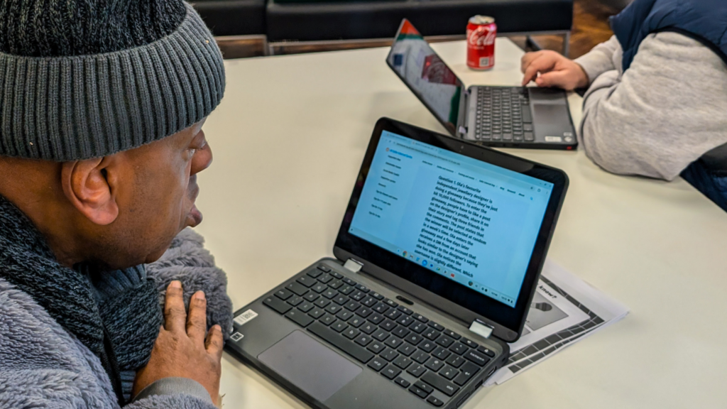 A person wearing a grey beanie and coat is seated at a table, looking at a Chromebook screen displaying a document with text. Another participant, wearing a blue vest, is using a laptop in the background. A can of Coca-Cola sits on the table, along with printed worksheets. The setting appears to be a group learning or training session.