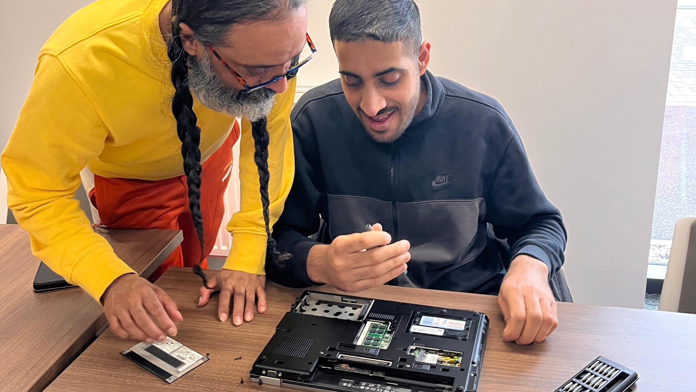 two people working together on a laptop repair. One person, wearing a yellow sweatshirt and glasses, is leaning over to assist the other person, who is in a black jacket and smiling while holding a small tool. The laptop is open on a wooden table, with its internal components exposed, and a small screwdriver set nearby. They appear engaged and focused on the task.