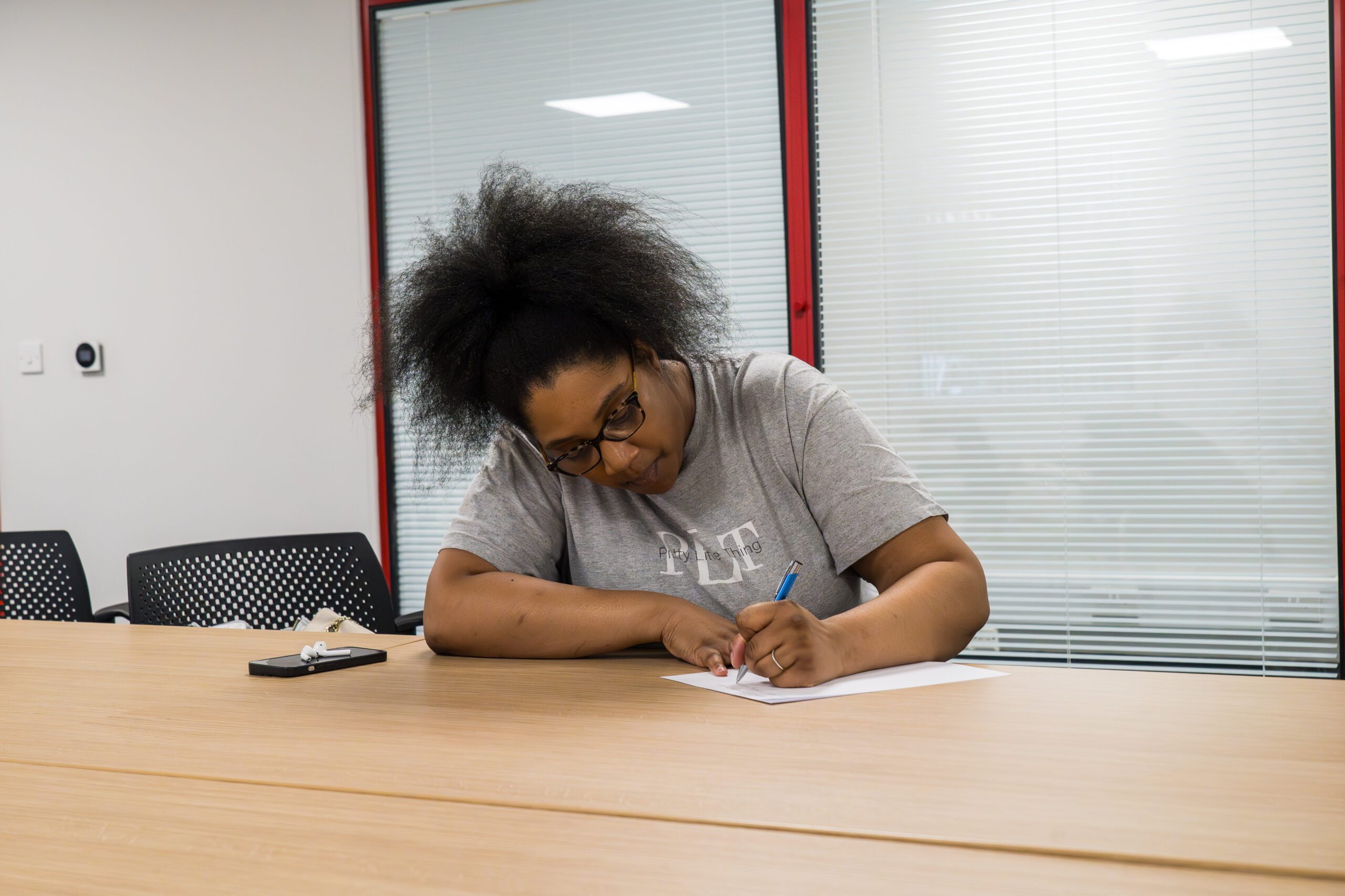 A woman with glasses and natural hair sits at a table, writing on a sheet of paper with a pen. She is wearing a grey t-shirt with text that appears to say "PIP Learning." A smartphone with white earphones is on the table next to her.
