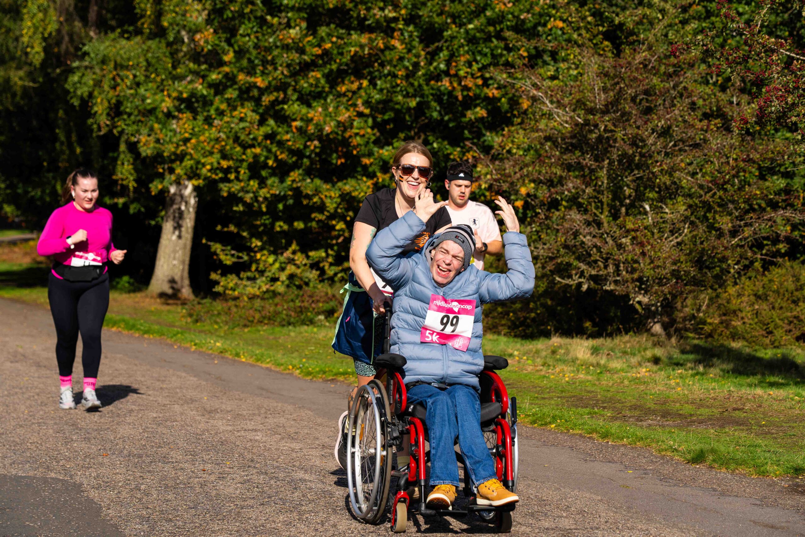 A smiling man in a wheelchair takes part in a 5K race in a park. He wears a blue jacket and a race number 99 with a pink “5K” and Midland Mencap logo. A woman runs behind him, pushing his wheelchair. Two other runners follow on the path. The sun is shining and there are green trees in the background.
