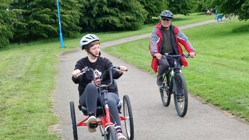 A young person wearing a helmet rides a red recumbent tricycle along a park path. Behind them, a man in a red jacket and helmet rides a bicycle. The path winds through a green park with trees and grass on a cloudy day.