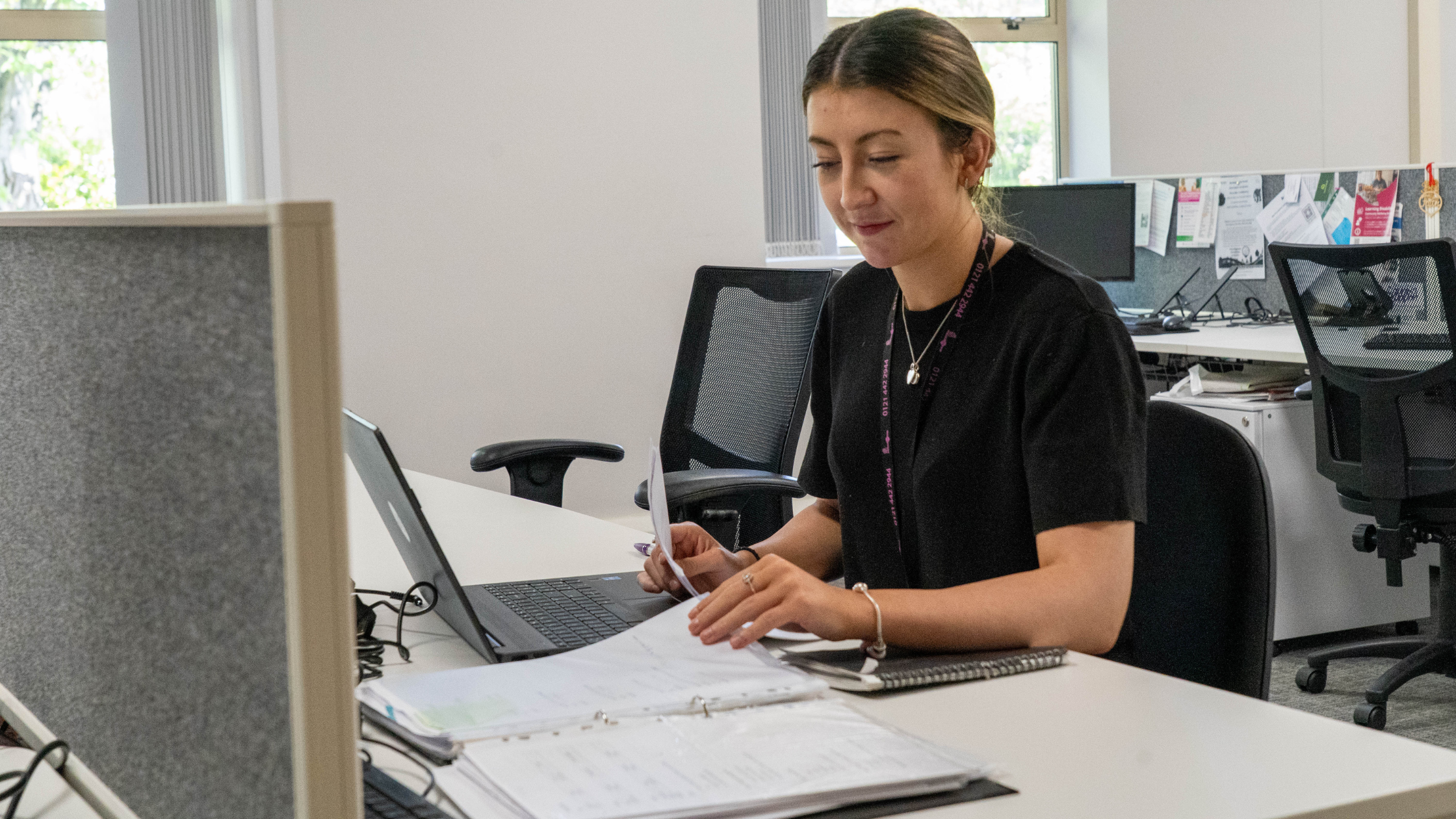 A woman sits at a desk in an office, looking at paperwork beside an open laptop. She is wearing a black top and a staff lanyard. Papers, folders and a notebook are spread across the desk, with office chairs, computer screens and noticeboards visible in the background.