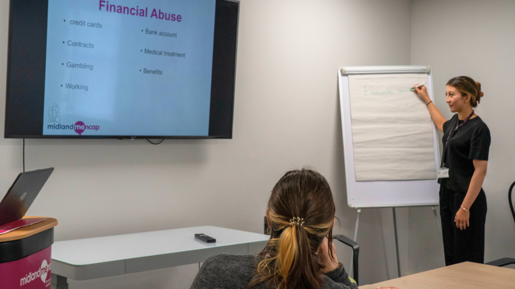 A staff member stands beside a flip chart, leading a training session about financial abuse. A presentation screen displays the heading “Financial Abuse” with topics including credit cards, contracts, gambling, bank accounts, medical treatment and benefits. One person is seated in the audience facing the presentation.