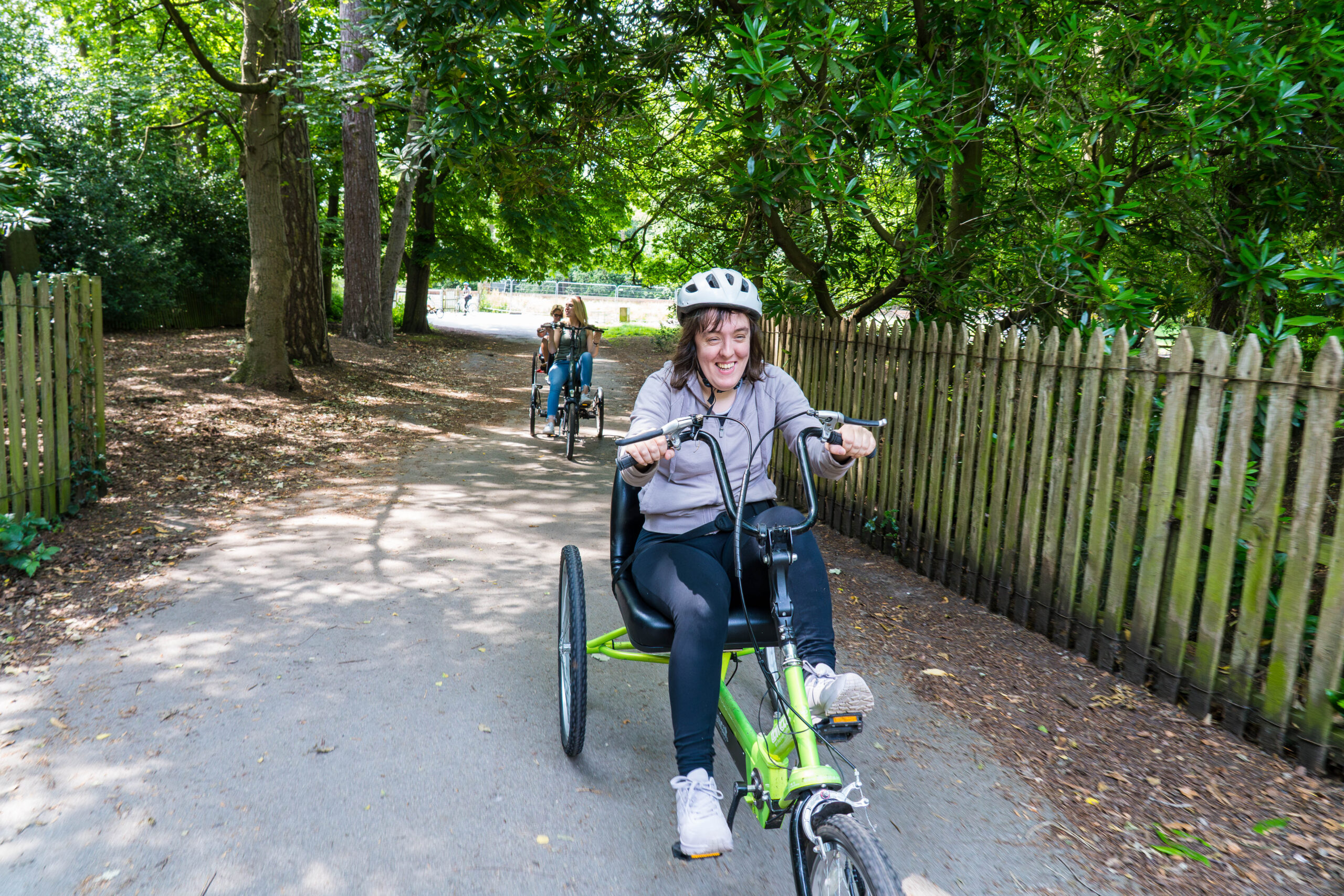 A smiling woman wearing a helmet rides a green adapted tricycle along a tree-lined park path. Two other cyclists follow behind her on adapted bikes.