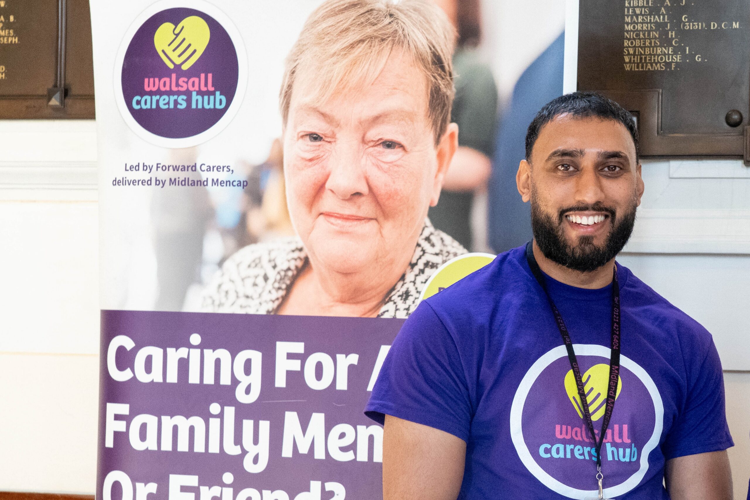 A smiling Walsall Carers Hub staff member wearing a purple branded T-shirt stands in front of a Walsall Carers Hub banner. The banner says the service is led by Forward Carers and delivered by Midland Mencap.
