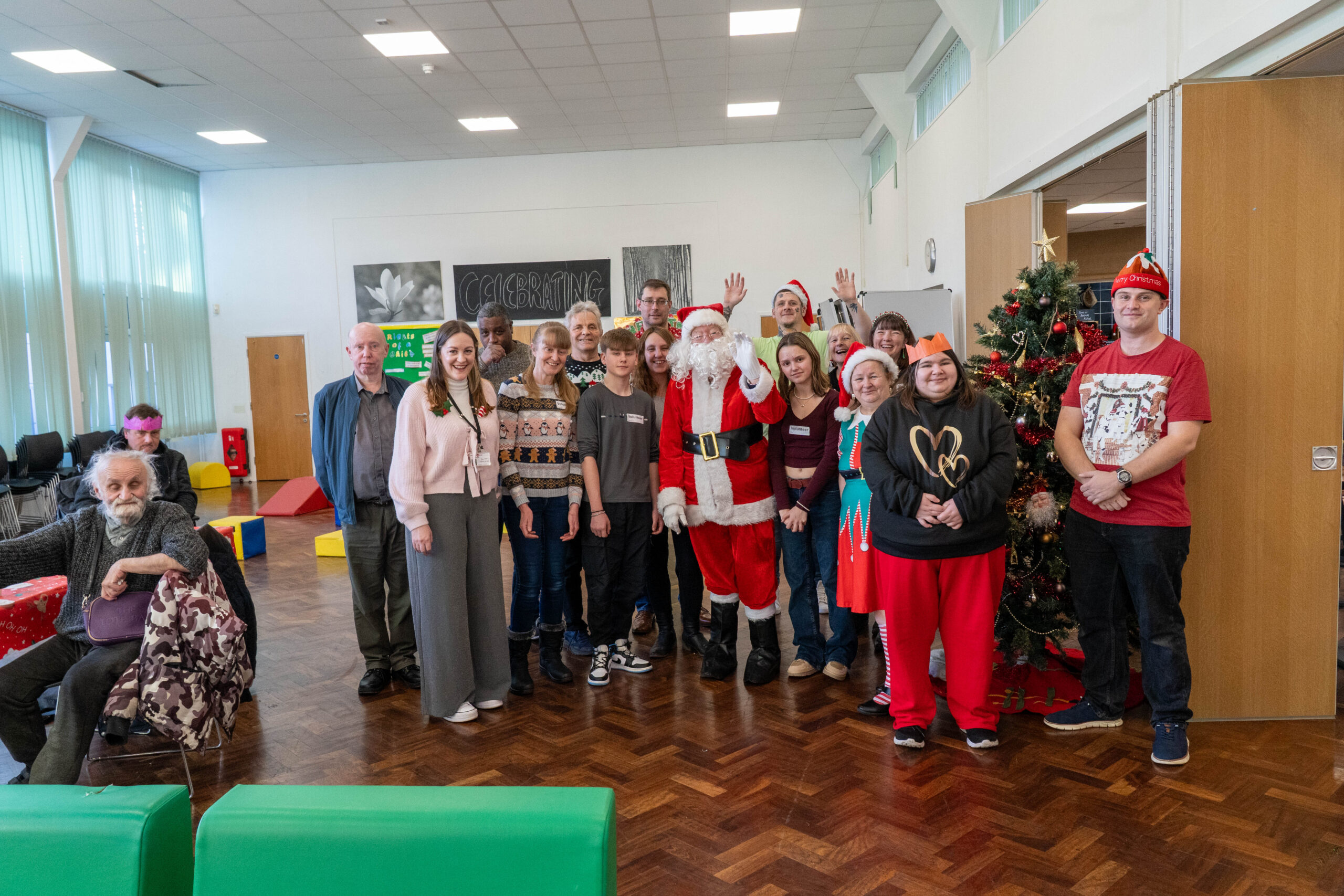 A group of people pose for a Christmas photo in a community hall. One person is dressed as Santa, and others wear festive jumpers and hats. A decorated Christmas tree stands beside the group. Two people are seated to the left of the photo.